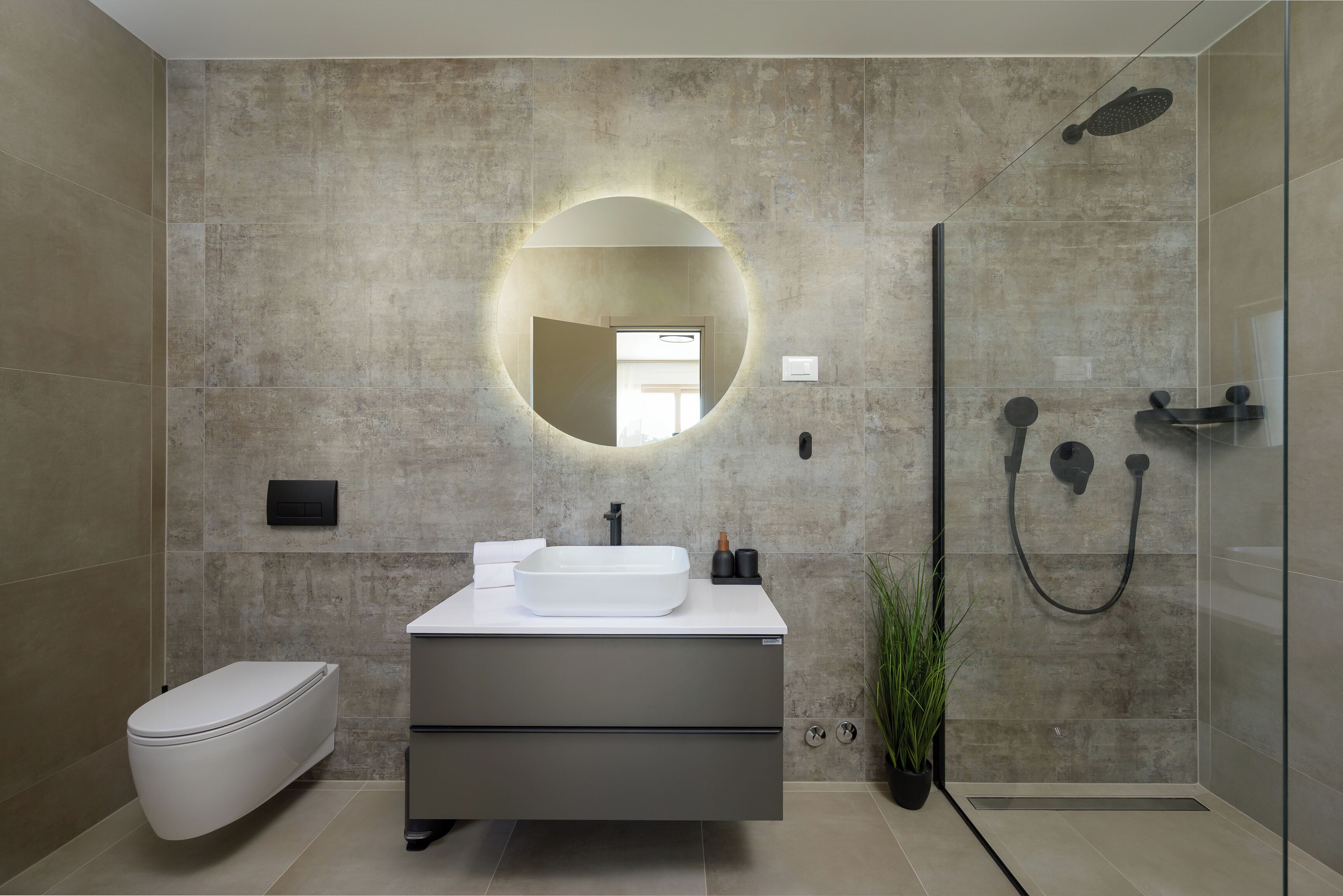 Une salle de bain moderne avec un grand miroir rond, un lavabo blanc, des toilettes et une douche à parois de verre.