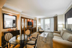 Ein Hotelzimmer mit Essbereich, einer Couch, einem Fernseher und einem großen Fenster mit Vorhängen.