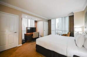Ein Hotelzimmer mit einem großen Bett, einem Schreibtisch mit Fernseher, einem Sessel und Blick auf die Stadt durch das Fenster.