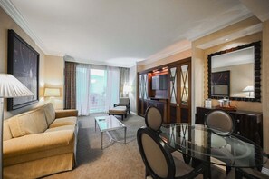 Ein Hotelzimmer mit einer Couch, einem Glastisch, einem Esstisch mit Stühlen, einem Fernseher und einem großen Fenster mit Vorhängen.