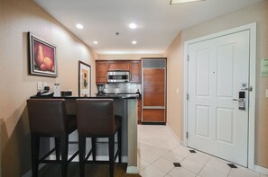 A compact kitchen with a microwave, oven, and refrigerator. There are two bar stools and a small island with a sink. The room has beige walls and a white door with a lock. - SVS: One Bedroom | Strip View Balcony | MGM Signature (Las Vegas)