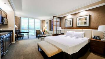 Quarto de hotel com cama, escrivaninha, cadeira, banqueta, cozinha compacta e uma janela grande com vista.