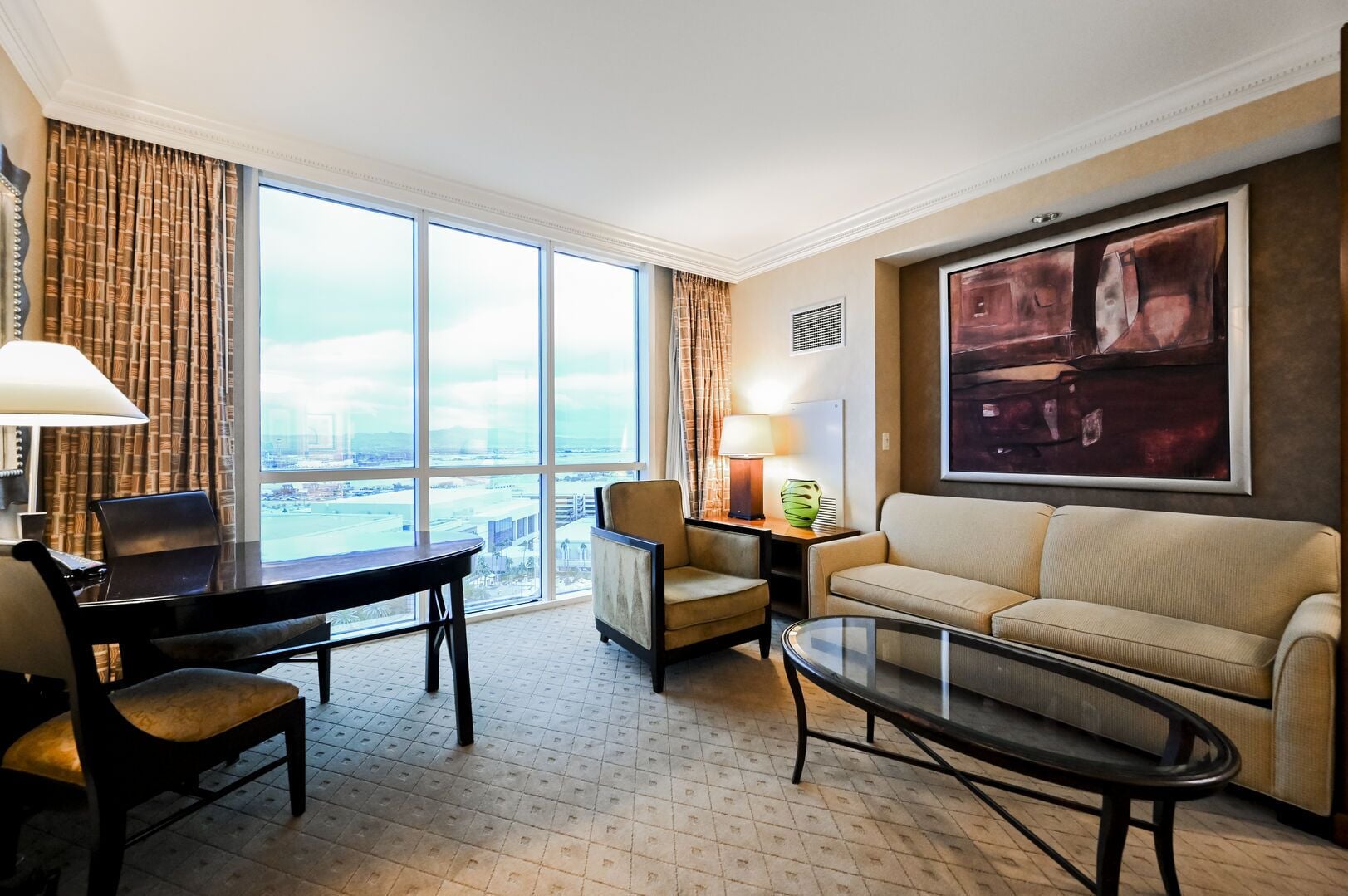 Quarto de hotel com uma janela grande, uma escrivaninha com cadeira, um sofá, uma mesa de centro e um abajur.