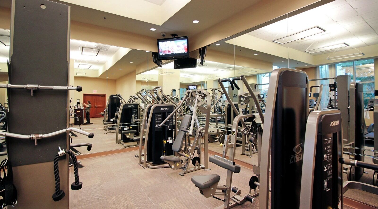 Ein Fitnessraum mit verschiedenen Trainingsgeräten, einem an der Decke befestigten Fernseher und einem Spiegel an der Wand.
