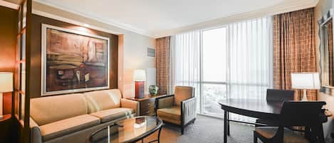 Een hotelkamer met een bank, een kleine tafel, een eettafel met stoelen, een groot raam met gordijnen en een schilderij aan de muur.