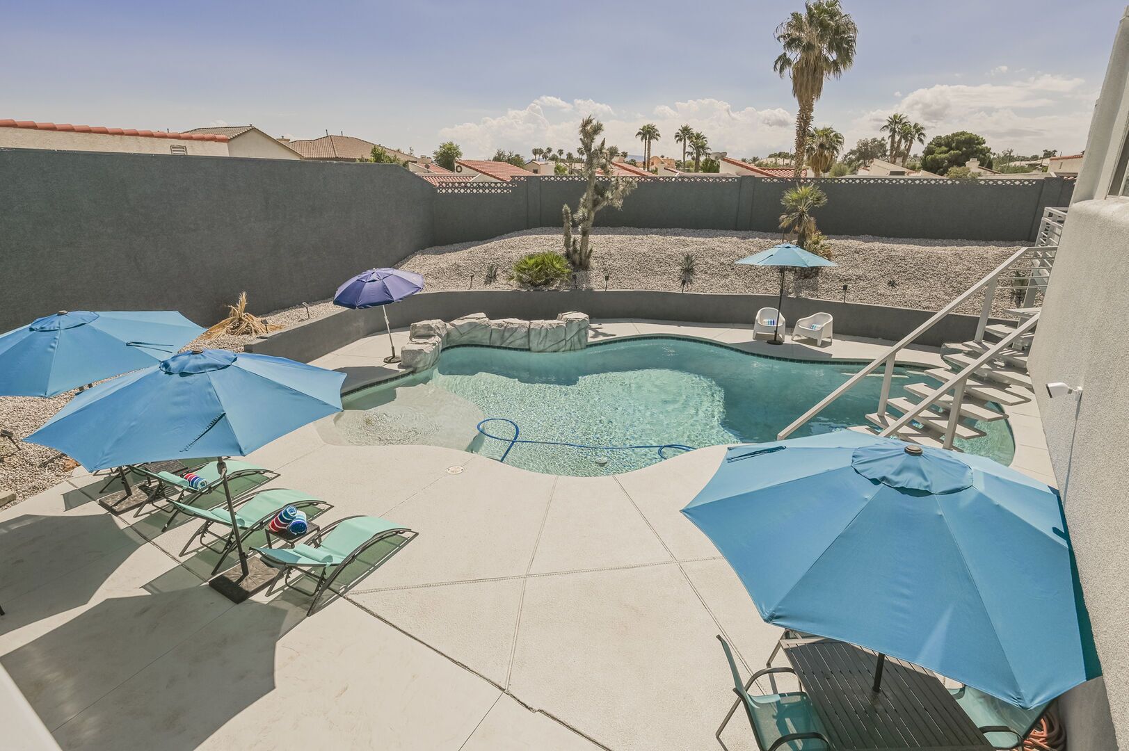 A swimming pool with lounge chairs and umbrellas, surrounded by a low wall and palm trees.