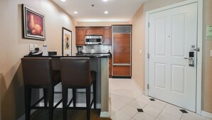 A compact kitchen with a microwave, oven, and refrigerator. There are two bar stools and a small island with a sink. The room has beige walls and a white door with a lock. - SVS: One Bedroom | Strip View Balcony | MGM Signature (Las Vegas)