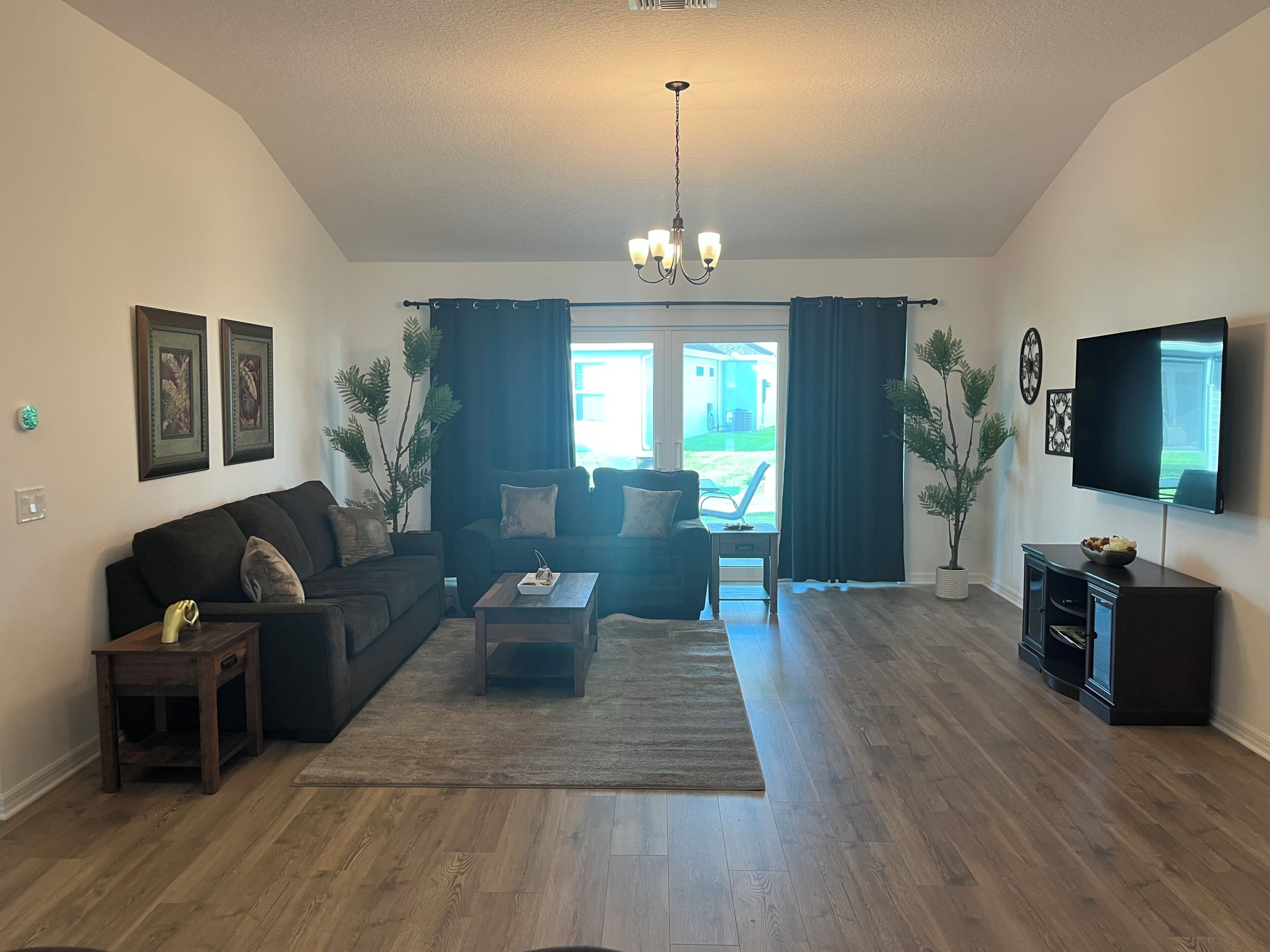 Ein Wohnzimmer mit einem dunkelgrauen Sofa, einem Couchtisch und einem Flachbildfernseher. Eine Kristallleuchter hängt von der Decke.