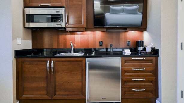 A compact kitchen with a microwave, a built-in TV above the counter, a stainless steel refrigerator, and wooden cabinetry. - SVS: Sphere Strip View Balcony | Signature at MGM (Las Vegas)