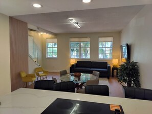 Un salon moderne avec une table en verre, un canapĂ© foncĂ©, des fauteuils jaunes et de grandes fenĂȘtres donnant sur un paysage verdoyant.