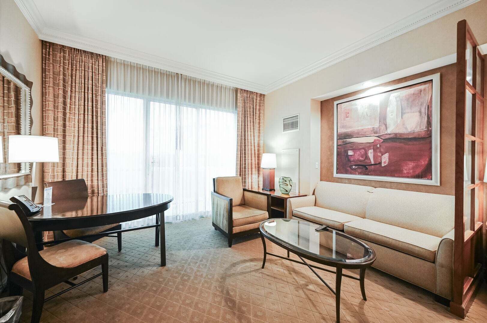 A hotel room with a dining table, two chairs, a sofa, a coffee table, a lamp, and a painting on the wall.