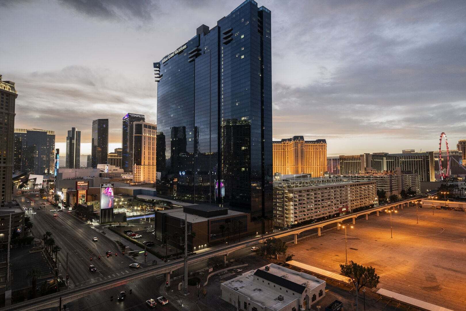 Un hotel de gran altura con fachada de vidrio moderna, situado en una zona urbana concurrida, rodeado de edificios altos y un amplio estacionamiento.
