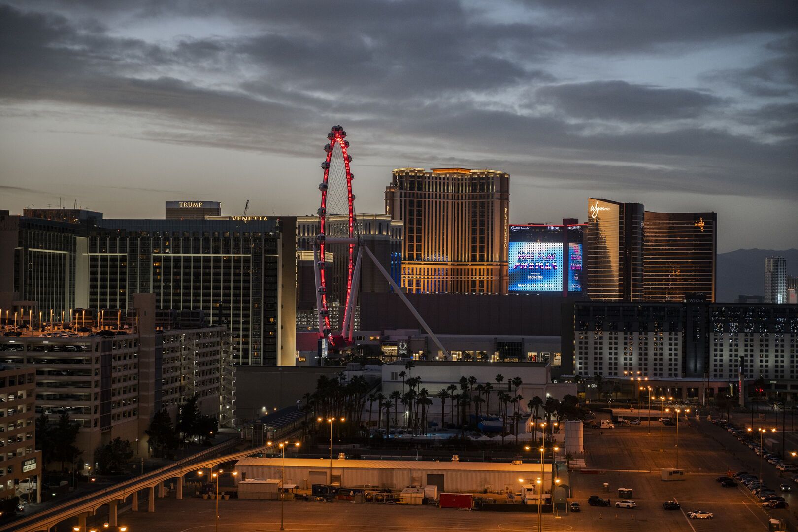 A cityscape with a prominent Ferris wheel, a large hotel with illuminated signage, and a parking area.