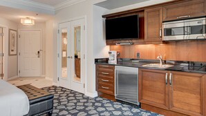 Ein Hotelzimmer mit Bett, Badezimmer mit Spiegel und Handtuchhalter, Küchenzeile mit Mikrowelle und Kaffeemaschine sowie ein Fernseher über dem Tresen.