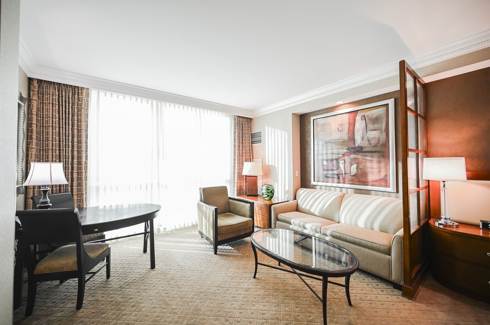 A hotel room with a dining table, a sofa, a chair, a glass coffee table, and a large window with curtains.