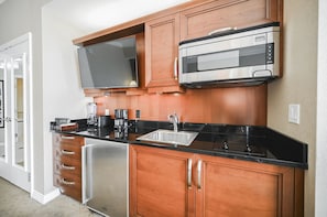 A kitchen with wooden cabinets, a stainless steel refrigerator, a black countertop, a built-in microwave, and a large flat-screen TV. - SVS: PH Floor Sphere Strip View Balcony | Signature at MGM (Las Vegas)