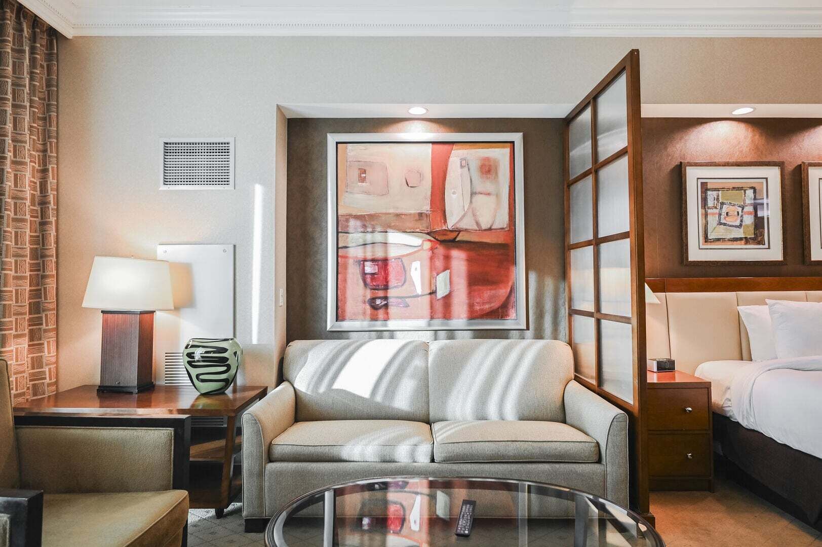 Une chambre d’hôtel avec un canapé, une table basse en verre, une lampe, un lit et des œuvres d’art accrochées aux murs.