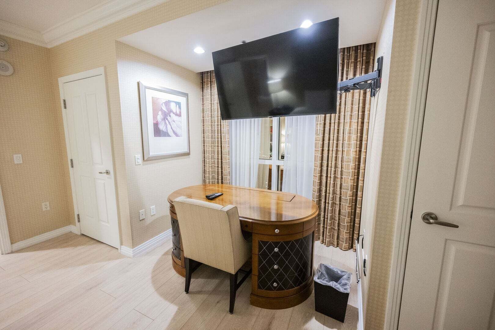 A hotel room with a round wooden table, a single chair, a television mounted on the wall, a framed picture on the wall, and a door.