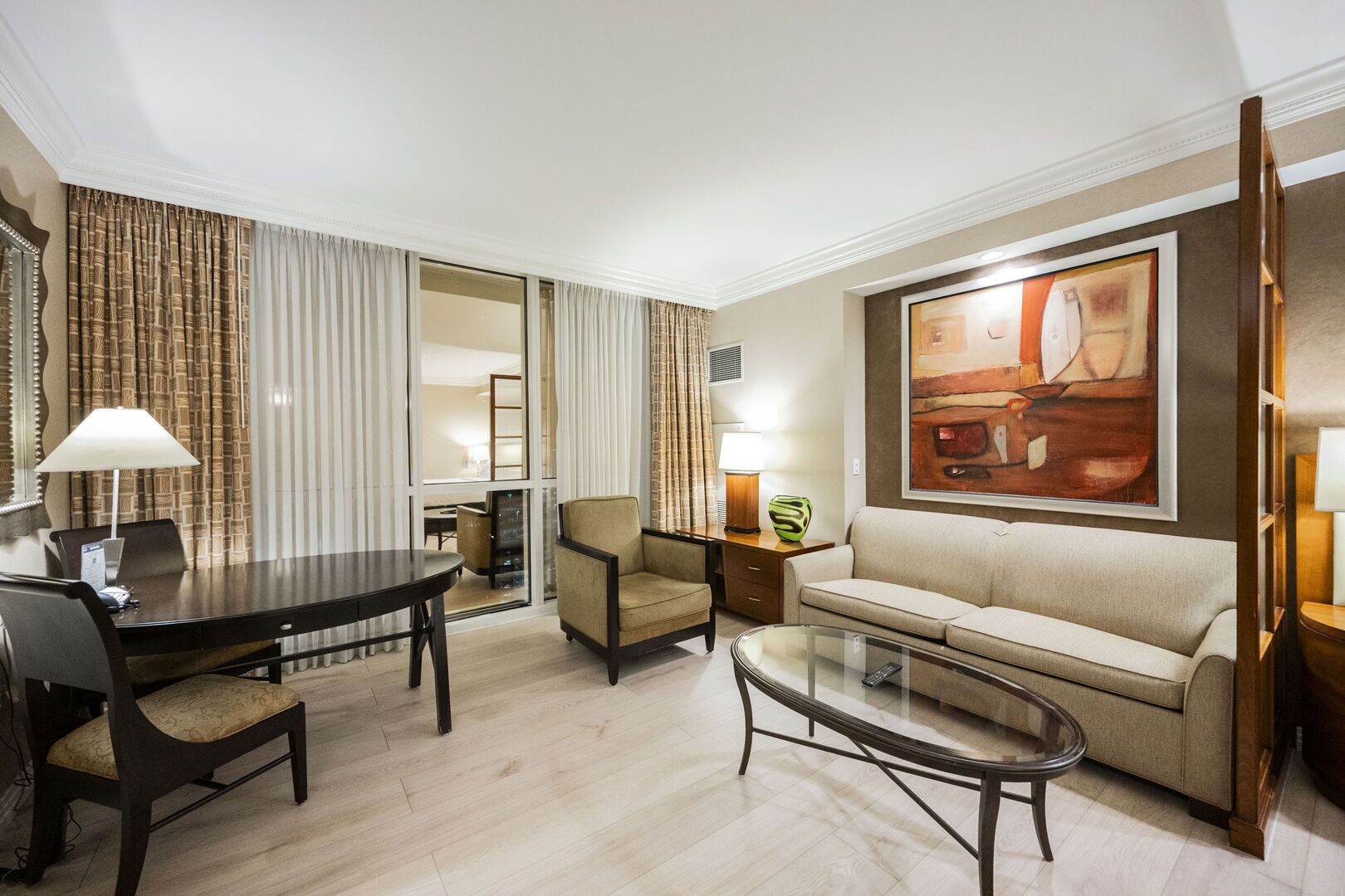 A hotel room with a dining table, two chairs, a sofa, a glass coffee table, and a painting on the wall.