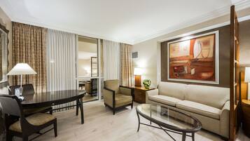 A hotel room with a dining table, two chairs, a sofa, a glass coffee table, and a painting on the wall.