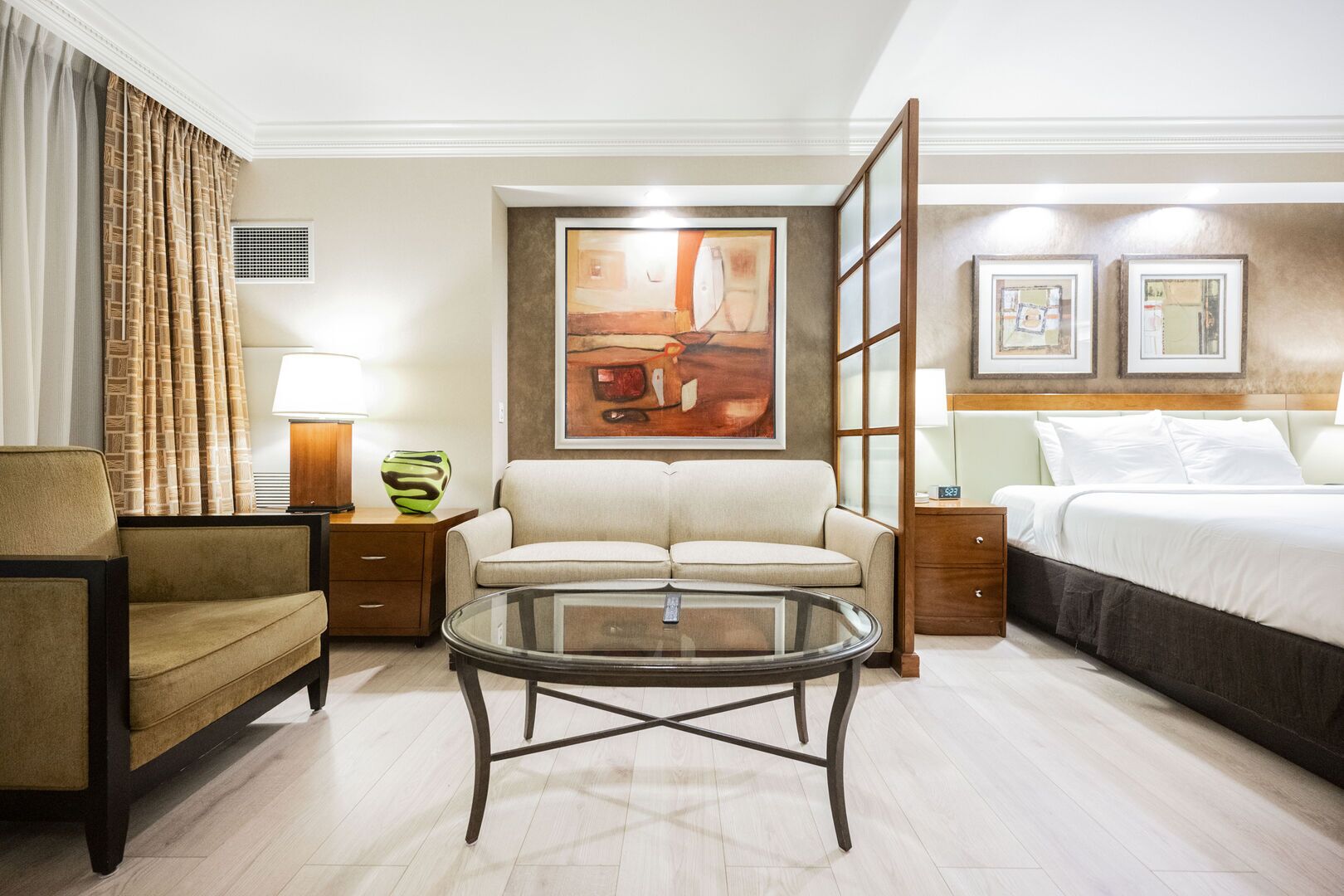 Quarto de hotel com sofá, mesinha de centro de vidro, cama, mesa de cabeceira com abajur e um quadro na parede.