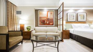 Quarto de hotel com sofá, mesinha de centro de vidro, cama, mesa de cabeceira com abajur e um quadro na parede.