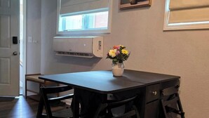 A room with a table, chairs, a vase with flowers, an air conditioning unit, and a window.