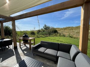 An outdoor patio with a wooden pergola, a black sofa, a table with a barbecue, and a view of a grassy field. - Duinvilla Wellness 5 (De Cocksdorp)