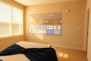 A hotel room with a bed, a wall-mounted screen displaying various streaming services, and a window with blinds.
