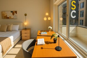 A modern hotel room with a bed, bedside table, lamp, desk, chair, and a large window with a view of buildings.