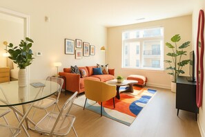 A modern living room with a glass dining table, orange sofa, and a large window with a view of buildings.
