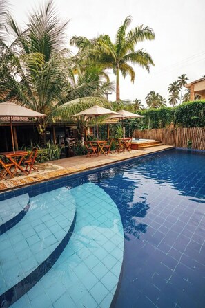 A swimming pool with lounge chairs and umbrellas, surrounded by palm trees and a wooden fence. - Jimbaran Pousada & Bistrot (Maragogi)