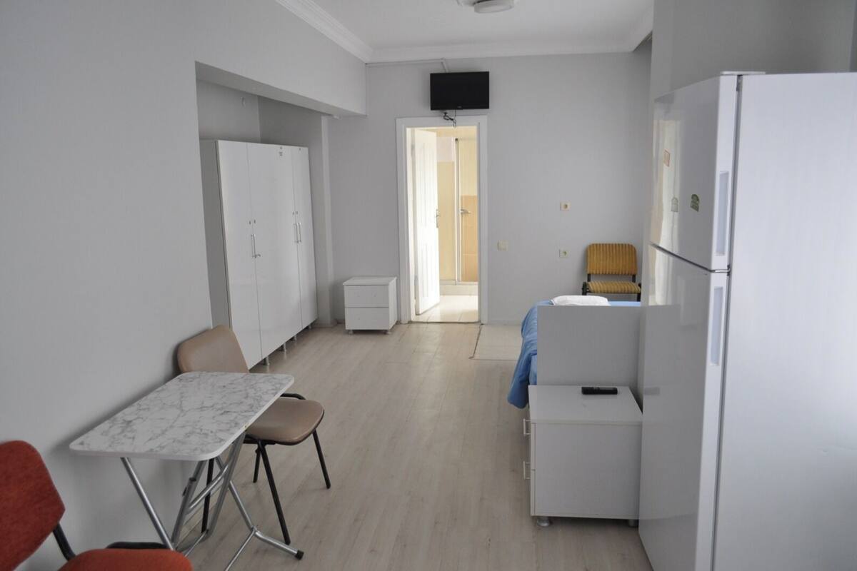 A small, clean room with a bed, a table, a chair, a wardrobe, a television, and a refrigerator.
