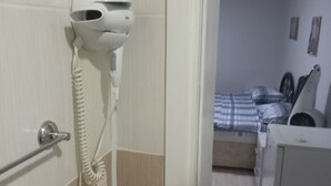 A hotel room with a hairdryer mounted on the wall, a bed with a checkered bedspread, a bedside table, and a small cabinet. - Bulut Gaye Pansiyon  (Edirne)