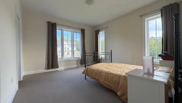 Ein Schlafzimmer mit Bett, Schreibtisch, zwei Fenstern mit Vorhängen und Blick auf ein Haus.