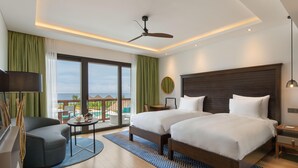 A hotel room with two beds, a desk, a chair, a balcony with a view, and a ceiling fan.
