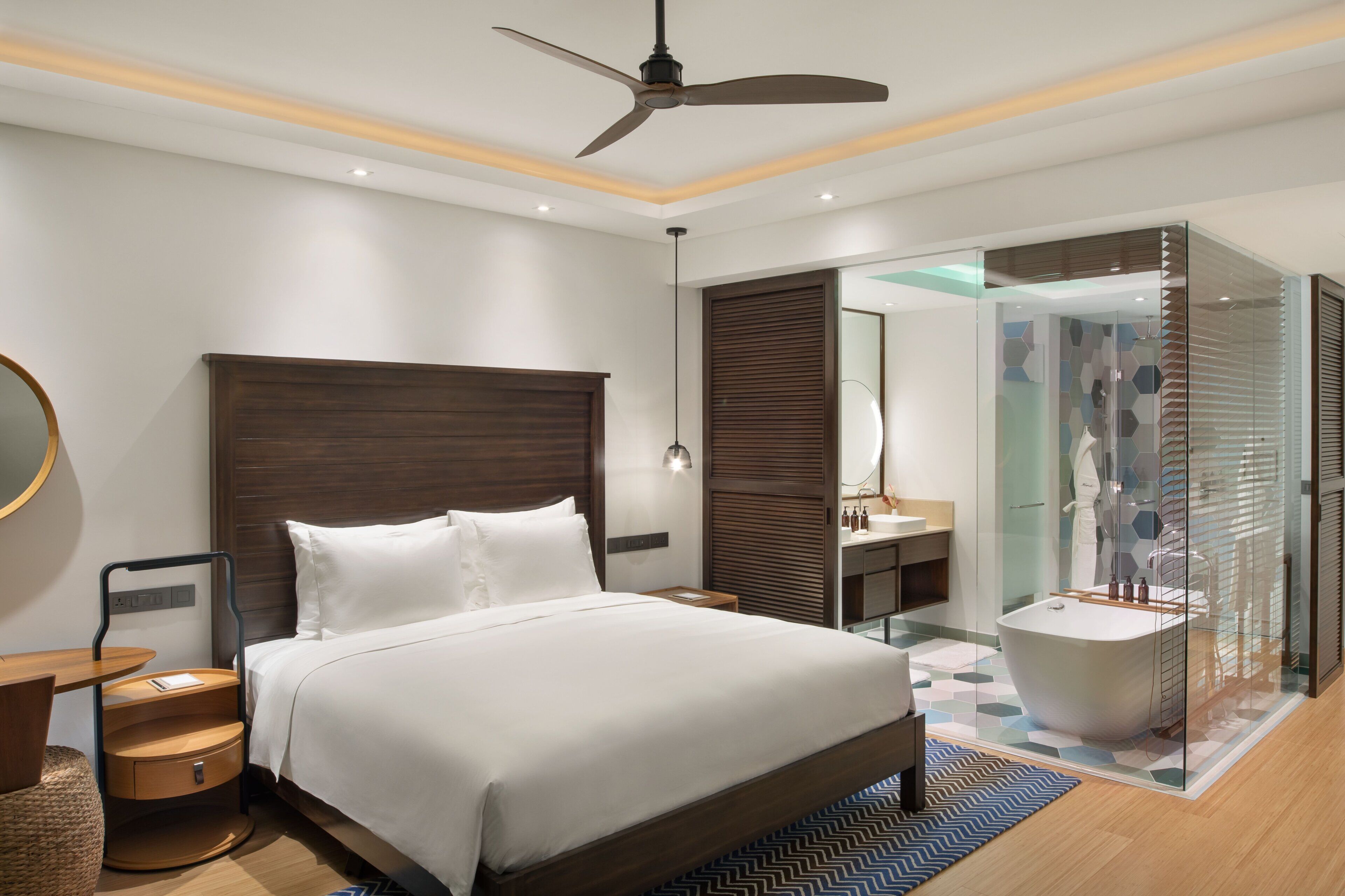 Ein modernes Hotelzimmer mit einem großen Bett, einem Deckenventilator, einem Badezimmer mit Duschkabine aus Glas und einem Holzboden.
