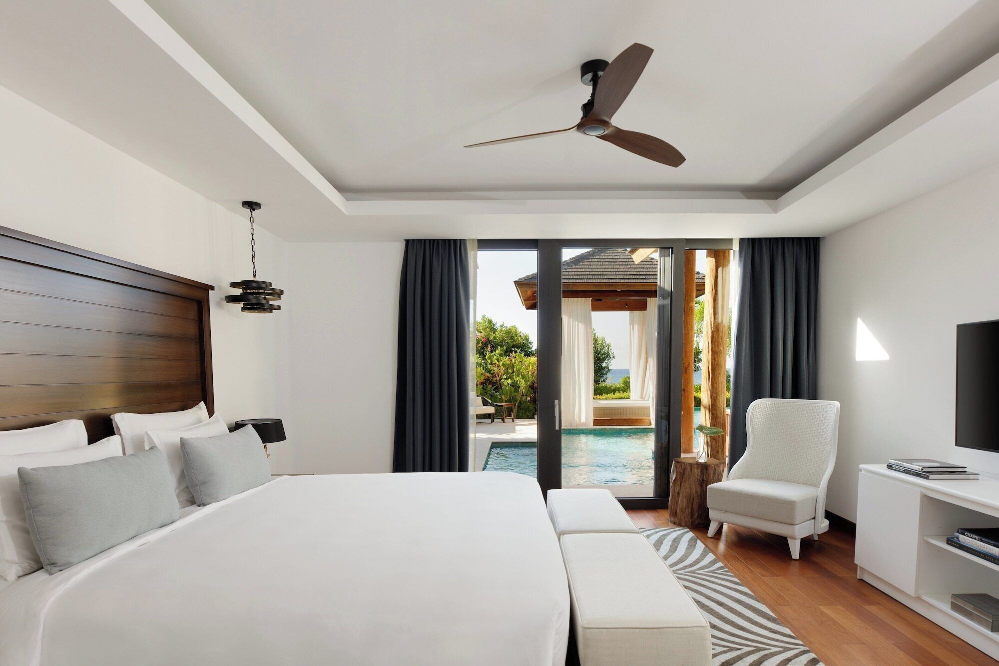 Ein modernes Hotelzimmer mit einem großen Bett, einem Deckenventilator, einem Fernseher, einem Schreibtisch und Blick auf einen Pool und einen Außenbereich.