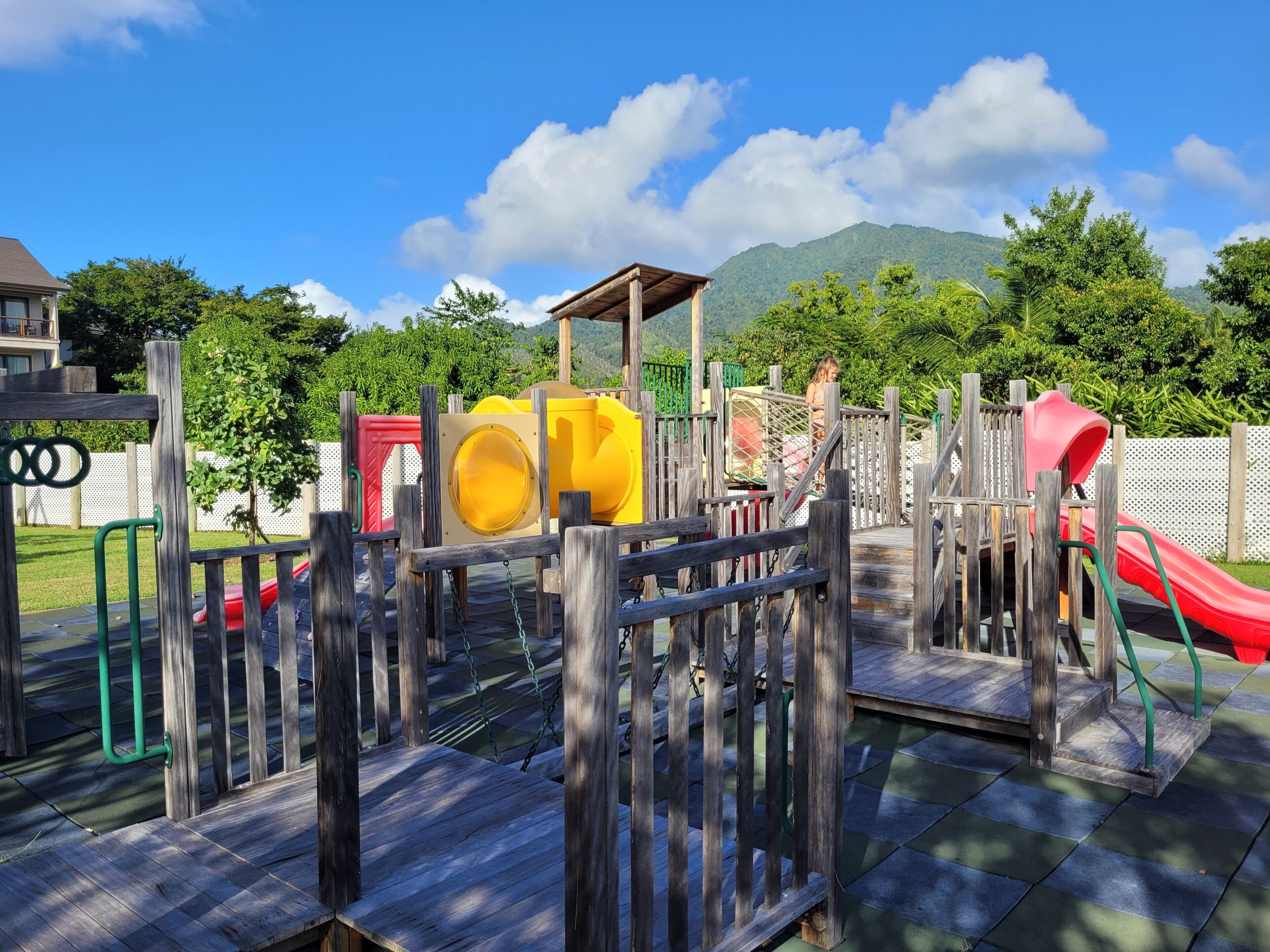 A playground with various play structures, including slides and climbing areas, surrounded by a wooden fence and greenery.