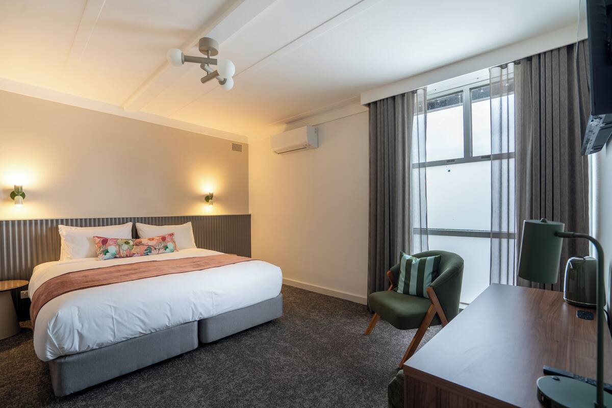 Ein Hotelzimmer mit Bett, Schreibtisch, Stuhl, Lampe und einem fensterbepflanzten Fenster.