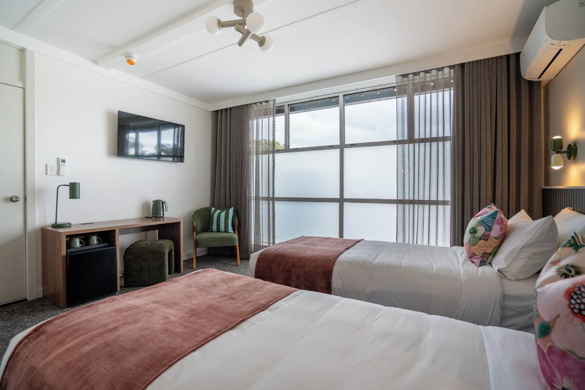 Ein Hotelzimmer mit zwei Betten, einem Schreibtisch, einem Sessel, einem Fernseher und einem großen Fenster.