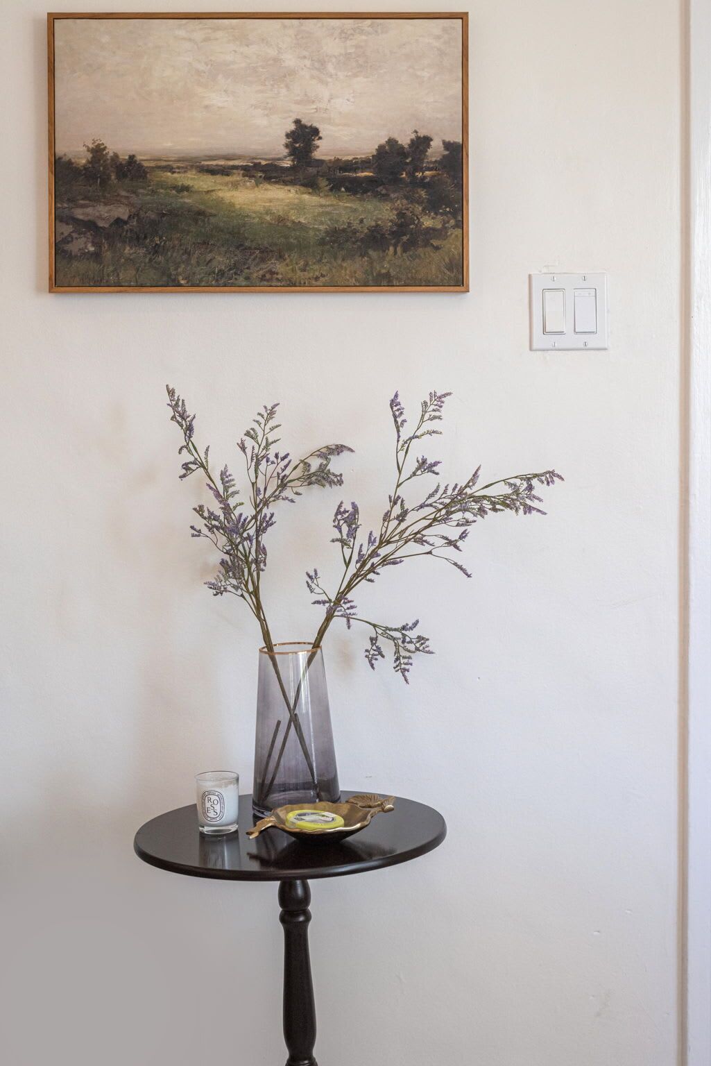 A vase with branches on a table, a candle, and a painting on the wall.