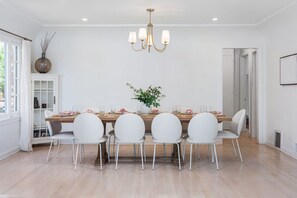 Comedor con una mesa de madera, sillas blancas, una lámpara de araña y una planta en maceta.