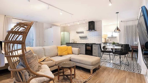 Een moderne woonkamer met een bank, een rieten stoel, een eethoek met tafel en stoelen, en een keuken met fornuis en kasten.