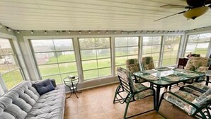 Une véranda avec un canapé confortable, une table en verre et des chaises, de grandes fenêtres et un ventilateur de plafond.