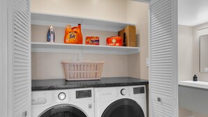 A laundry area with two washing machines, a basket, and shelves with laundry supplies. - Luz de Ventana | Redesigned 2BR Condo · Pools + Fitness · Mountain Views (Tucson)