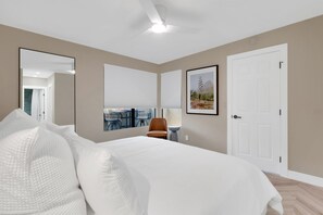 A bedroom with a bed, a chair, a desk, a window with blinds, a door, and a ceiling fan. - Luz de Ventana | Redesigned 2BR Condo · Pools + Fitness · Mountain Views (Tucson)