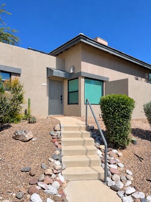 A modern building with a concrete staircase leading to a door, surrounded by rocks and small plants. - Luz de Ventana | Redesigned 2BR Condo · Pools + Fitness · Mountain Views (Tucson)
