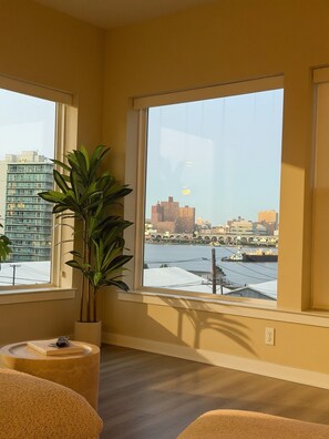 Ein Zimmer mit einem großen Fenster und Blick auf eine Stadtlandschaft und einen Fluss. Auf einem Beistelltisch steht ein Blumentopf.