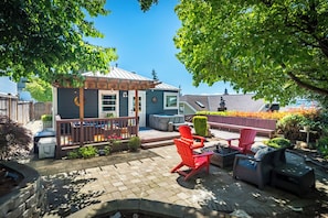 Eine Terrasse mit Sitzgelegenheiten im Freien, einer kleinen überdachten Konstruktion und Blick auf ein Haus im Hintergrund.